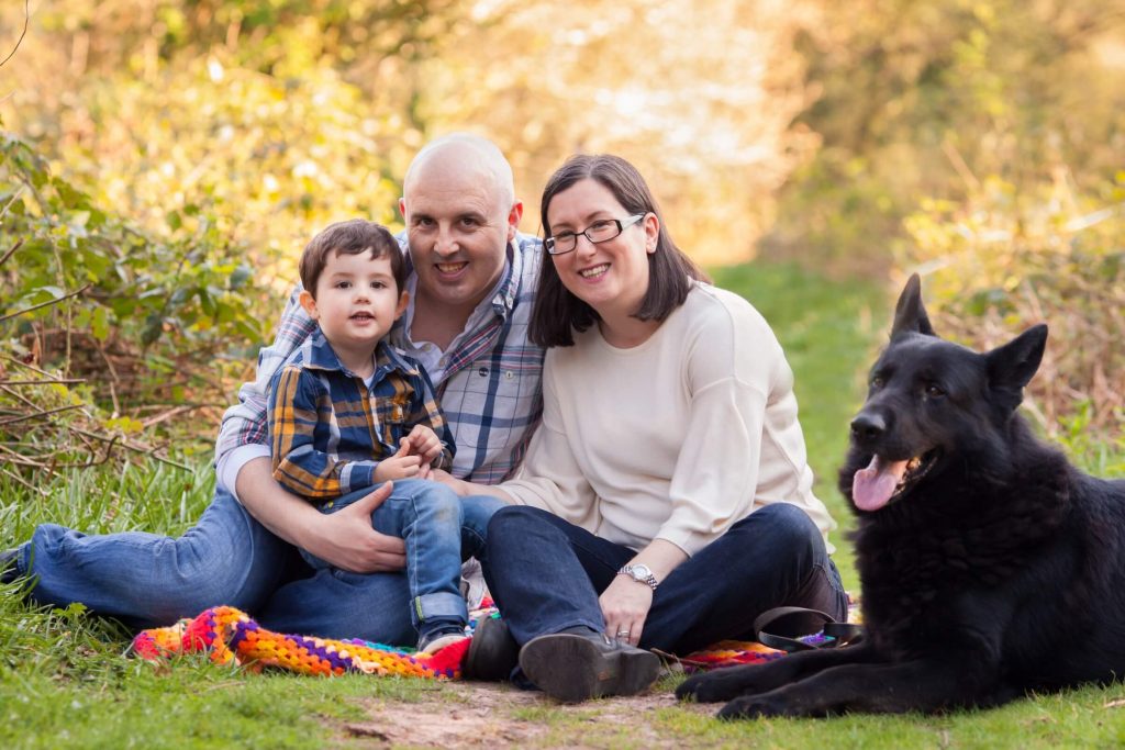 A picture of a family with the mu, dad and little boy sitting on a blanket in a woods with their black german shepherd dog beside them.