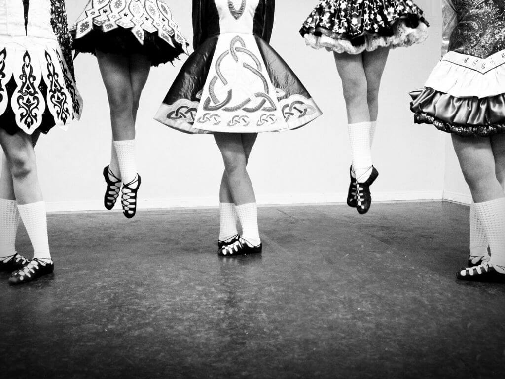 A black and white picture of the legs and feet of five Irish dancers in their dance shoes, socks and part of the skirt of their dresses showing