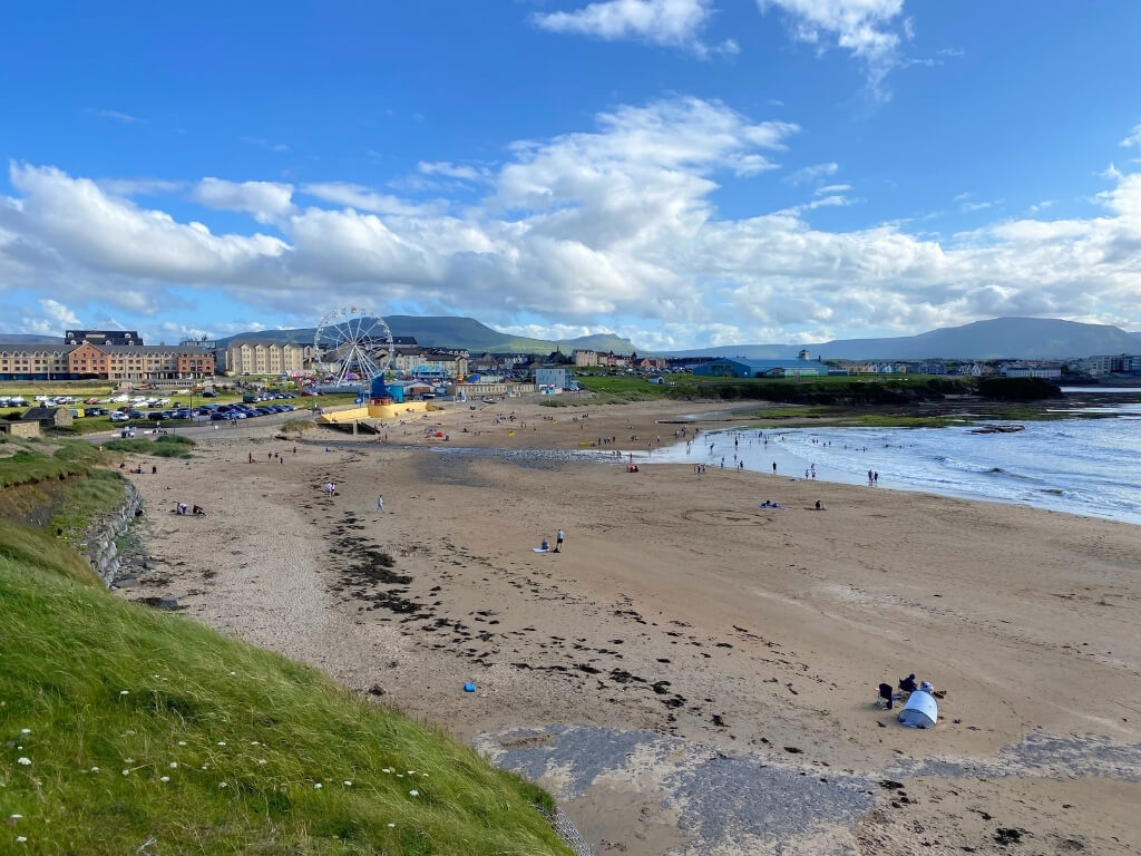 A picture of the golden sandy beach at Bundoran, Donegal with people on the beach and in the sea in the background. To the left is green hills and the ferris wheel at Bundoran and to the right is the white sea and mountains in the background