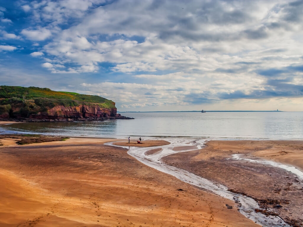 A picture of the sandy beach at Dunmore East with red cliffs on the left topped with green grass, a patchy blue sky in the background and a tidal stream of water in the foreground on the right, Three people are at the shoreline on the sand