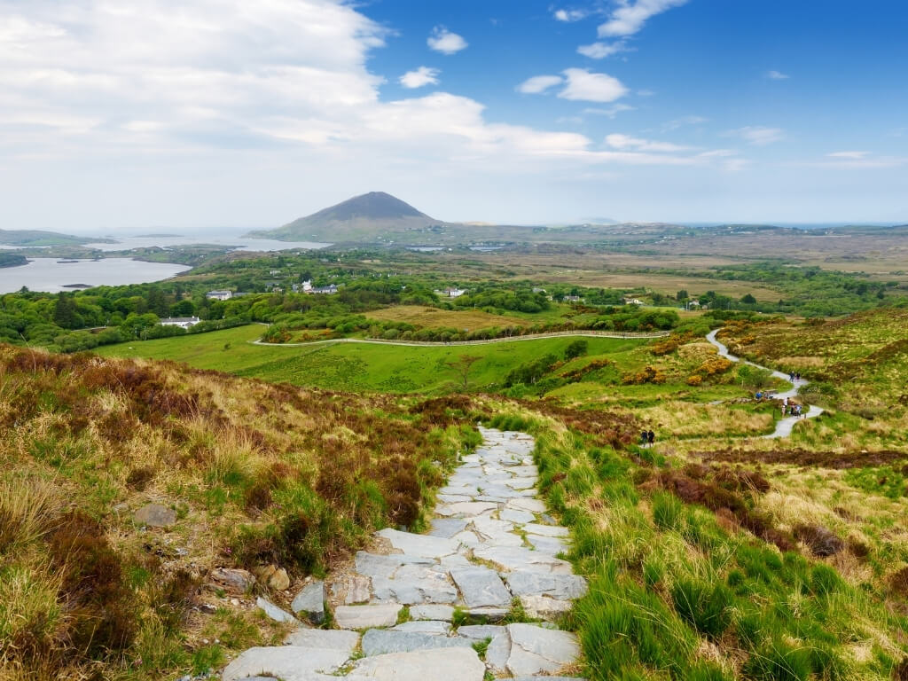 A view from the Diamond Hill Coastal Trail in Connemara National Park with a stoney path weaving its way through rolling fields and a sugar-loaf mountain in the background leading down to a lake