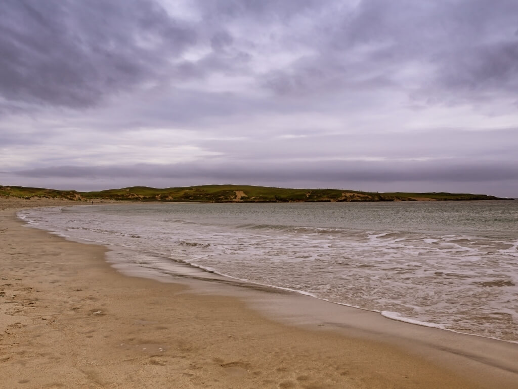 A picture of a grey, menacing sky overhead and the sandy beach of Dog's Bay in the foreground with green hills in the background