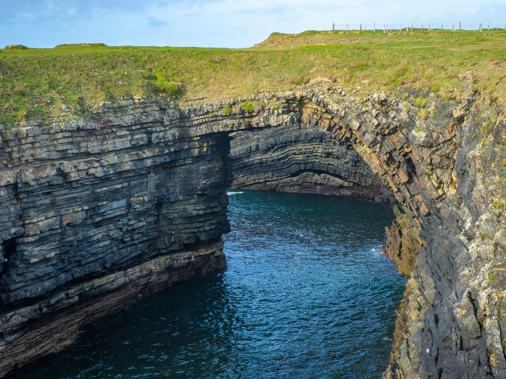 A picture of the Bridges of Ross on the Loop Head Peninsula in County Clare, Ireland, a sea bridge with green grass on top and dark blue sea waters flowing underneath it