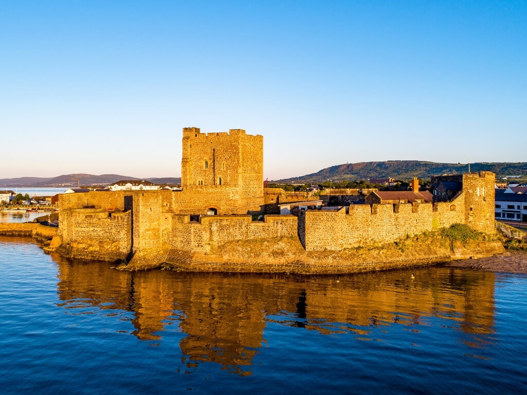 A picture of Carrickfergus Castle at the shoreline bathed in golden sunlight with blue skies overhead