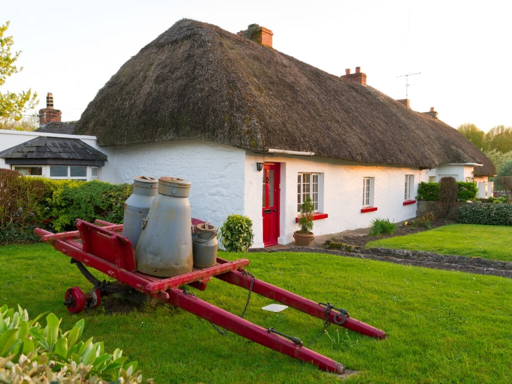 A picture of a traditional house with thatched roof in Ireland and an old red cart with old milk pails on it