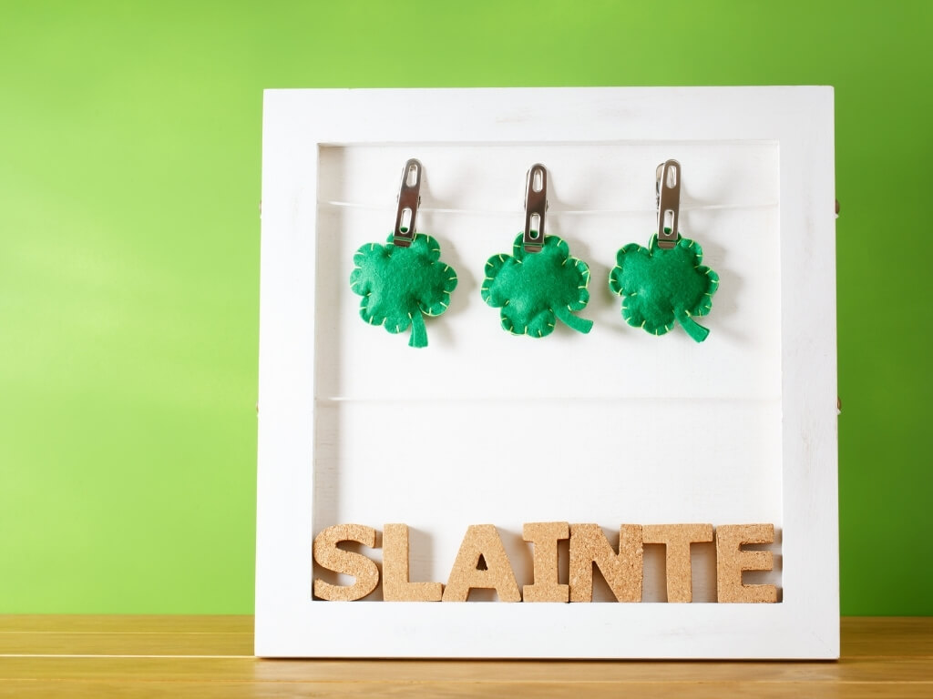 A picture of a white frame with stitched felt shamrocks on pegs at the top and cork letters on the bottom spelling out Slainte