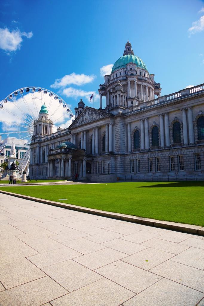 A picture of Belfast City Hall with a Ferris Wheel behind it, green grass in front and blue skies overhead