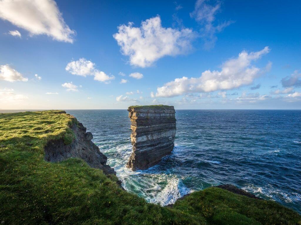 A picture of the sea stack of Downpatrick Head in County Mayo with waves crashing around it and blue skies overhead