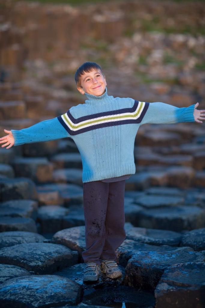 A picture of a boy in a blue woollen jumper and dark trousers standing at the Giant's Causeway, Northern Ireland with a smile on his face and arms open wide