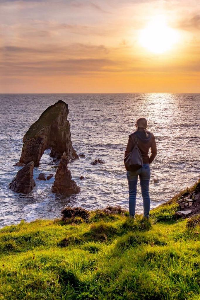 A woman in jeans and a jacket standing at the coast edge looking at the Crohy Sea Stacks in Ireland