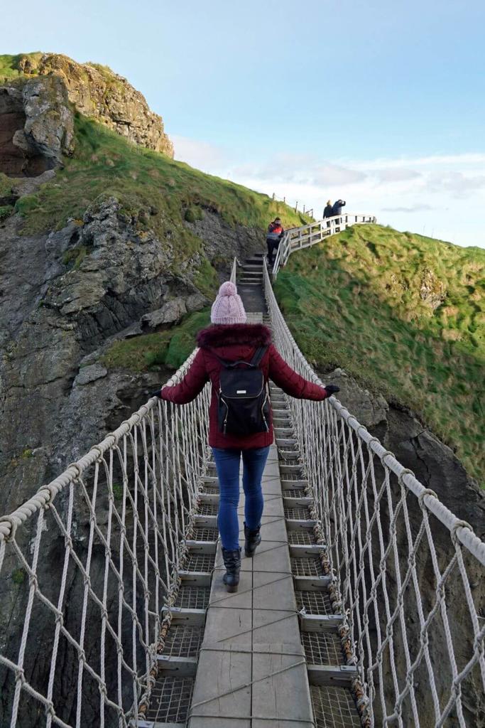 A picture of a woman in a wine coat and woollen hat crossing the Carrick-a-Rede Rope Bridge in Northern Ireland