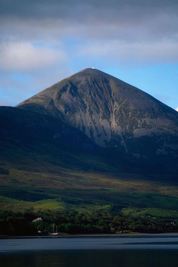 A picture of Croagh Patrick Mountain in County Mayo with the white church at the summit just visible, blue skies overhead and water below the mountain in the foreground with green fields slopping gently down to it