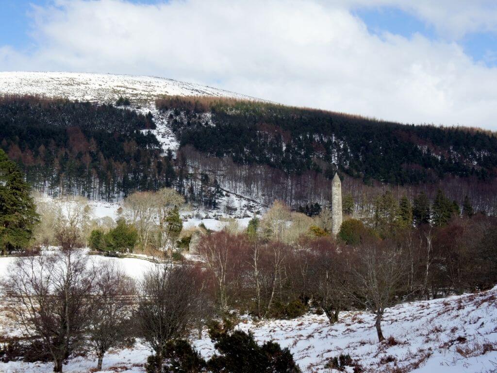 A picture of the Glendalough monastic site in winter with snow on the mountains behind and the hills in the foreground
