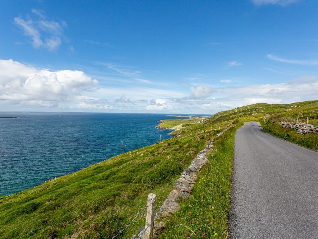 A picture of a road with grassy fields on the right and the blue waters of the Atlantic Ocean on the left along the Sky Road, Wild Atlantic Way, Galway
