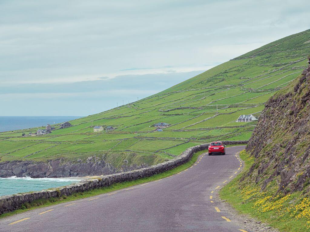 A picture of a red car on a narrow road in Ireland with the coast to the left of the road and hills to the right