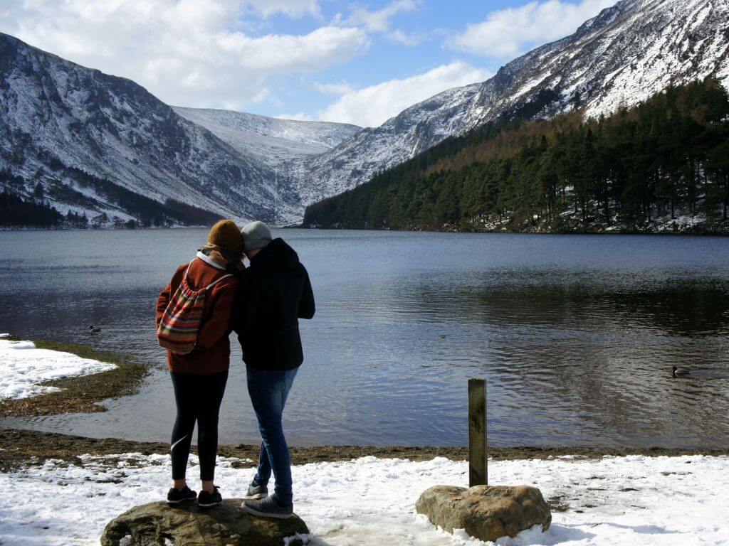 A picture of Glendalough's Upper Lake during winter with snow on the ground and mountains and two ladies standing in front of the lake