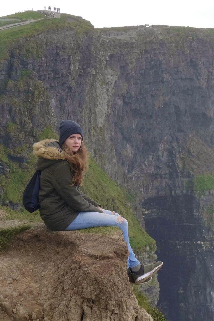 A lady in a hat and green winter coat sitting on top of the Cliffs of Moher