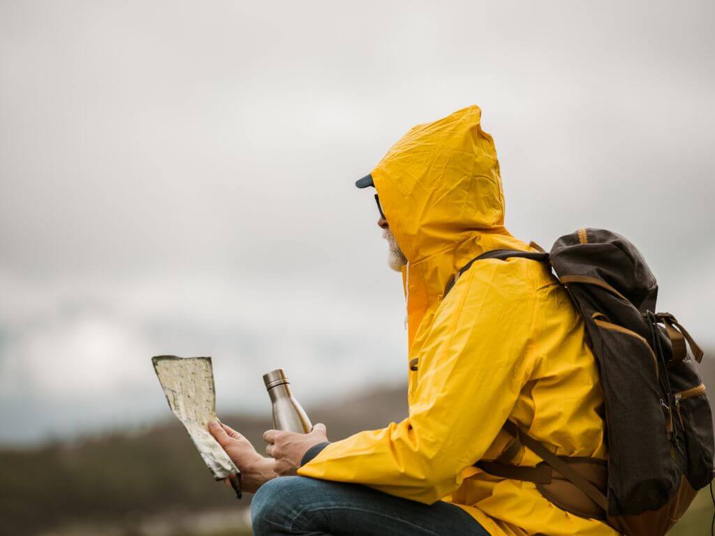 A picture of a man sat down with a flask in one hand, a map in the other and wearing a backpack and yellow raincoat