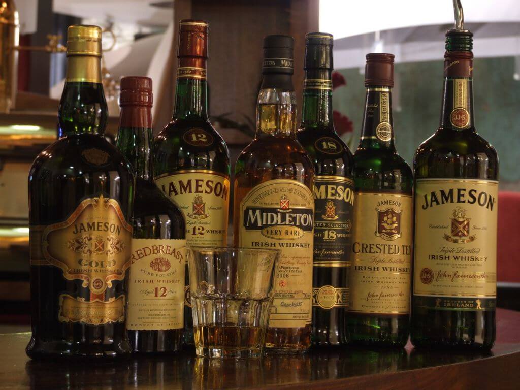 A picture of an assortment of bottles of Jameson Whiskey standing side by side on a bar with a glass in front of them containing a measure of whiskey