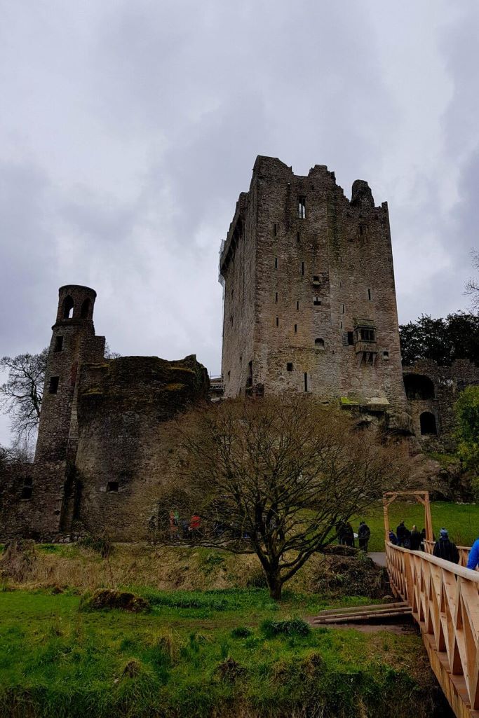 A portrait picture of Blarney Castle from the wooden bridge that approaches it and grey skies overhead