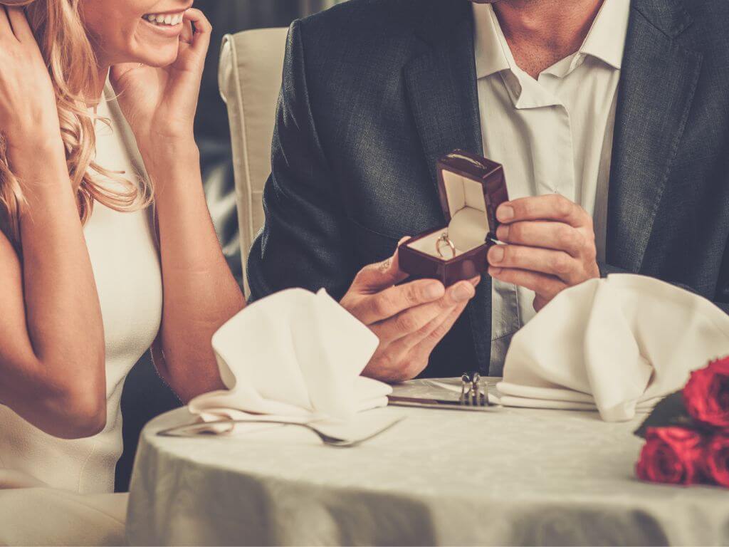 A piture of a man proposing marriage to a woman at a dinner table with an engagement ring in a jewelry box