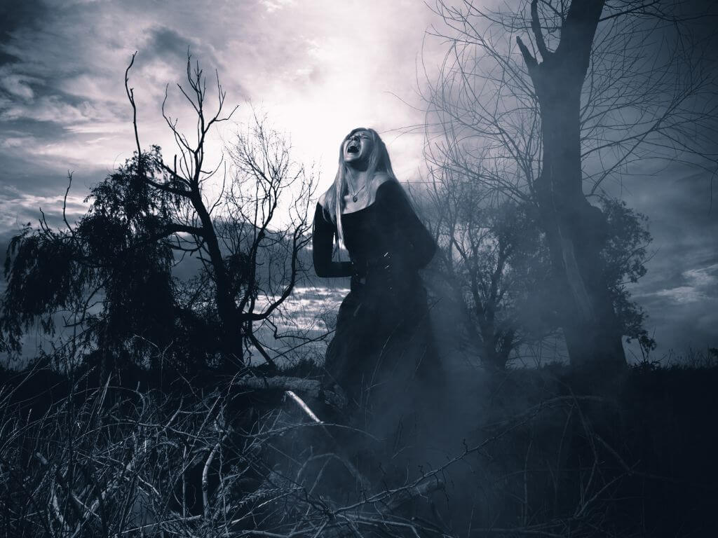 A black and white picture with a woman standing among trees and bushes looking like she is screaming to depict the image of a banshee wailing