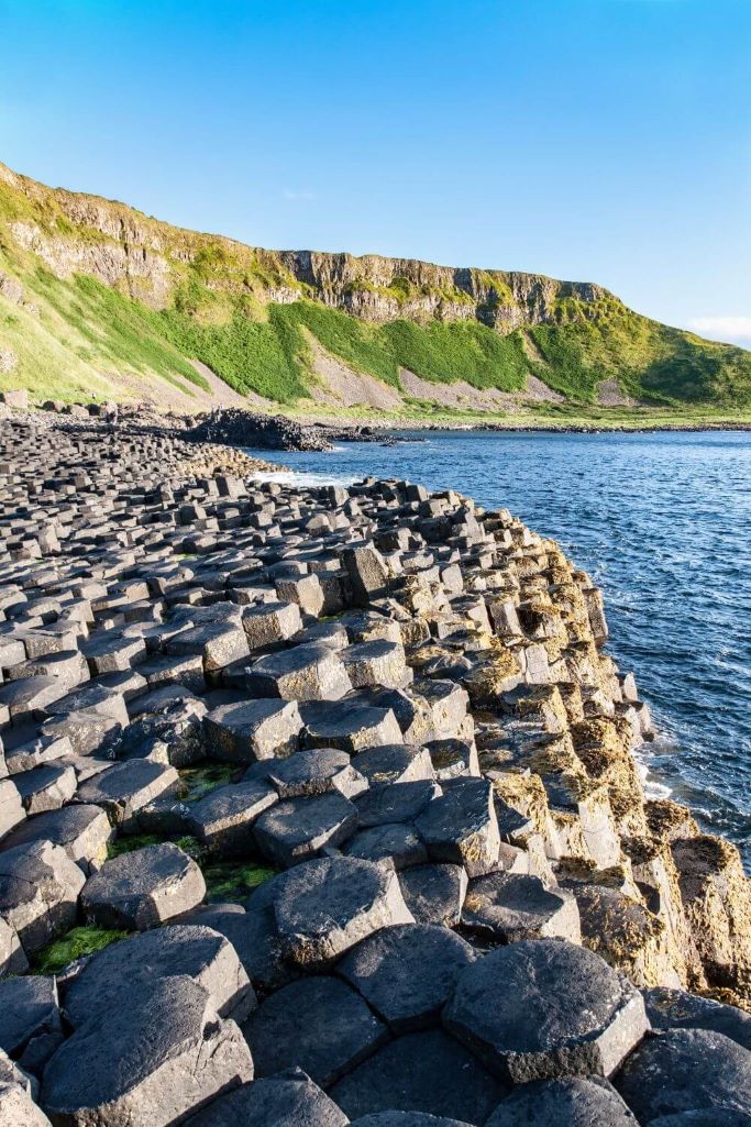 A picture of the Giant's Causeway in the foreground, green cliffs in the middle ground and blue skies overhead in the background