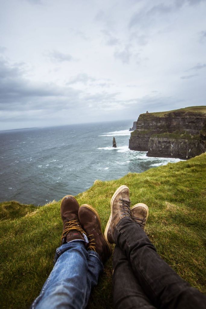 A picture of the legs and feet of two hikers sitting on the grass at the Cliffs of Moher, with part of the Cliffs visible in the middle ground and cloudy grey skies in the background