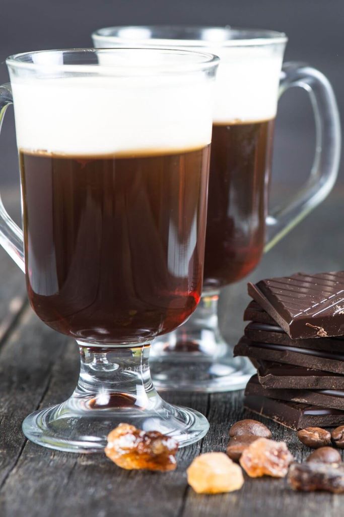A picture of two glass mugs of Irish Coffee on a neutral background with some brown sugar lumps and squares of chocolate to the right of the glass mugs