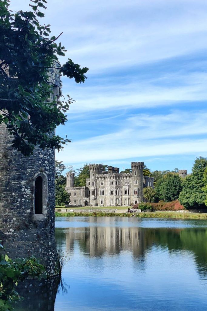 A picture of Johnstown Castle, Wexford from across the lake that sits in front of the castle, with a lake tower to the left of the picture and blue skies overhead