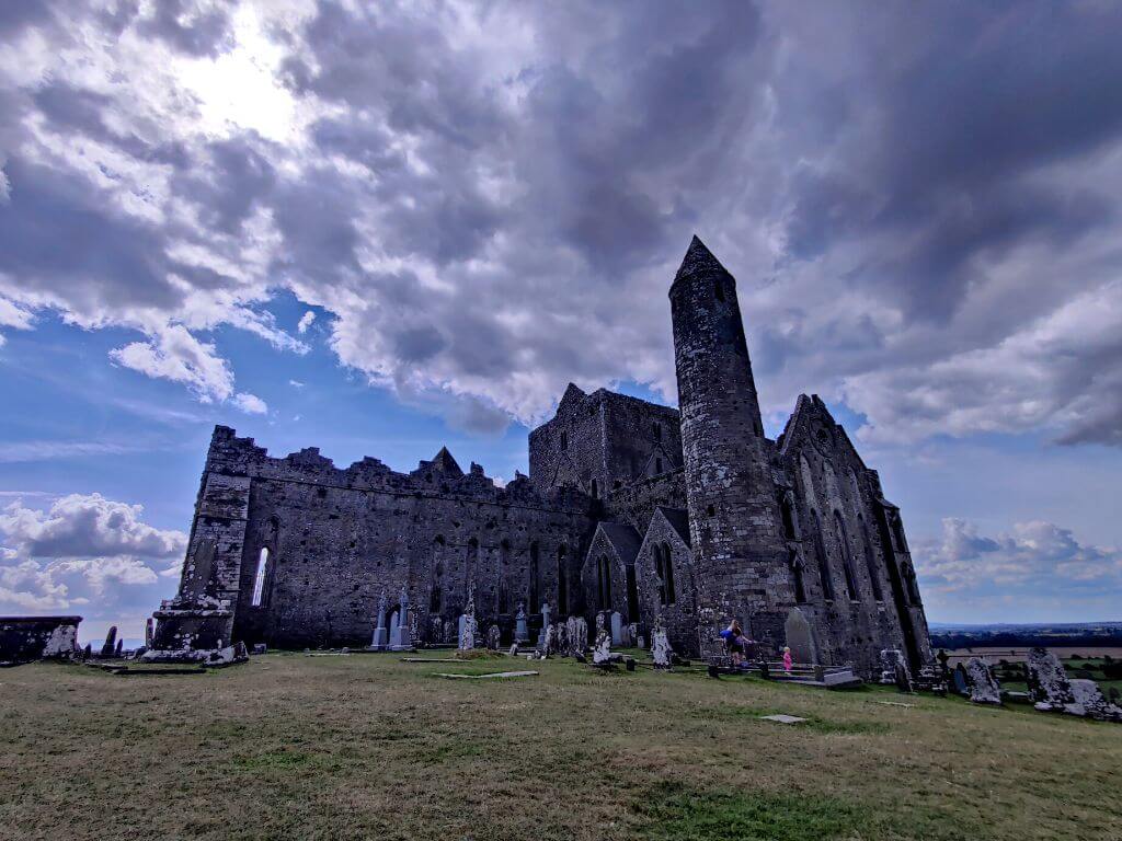 A picture of the Round Tower and Abbey of the Rock of Cashel in County Tipperary with green grass in the graveyard in front and blue skies overhead with some fluffy white clouds in the sky