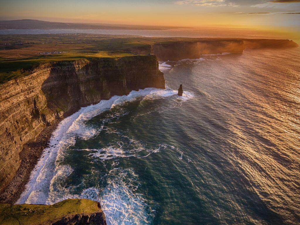 An aerial picture of the Cliffs of Moher from above at sunset with golden light lighting up the right hand top corner of the picture and white waves crashing beneath the cliffs on the left hand side of the picture