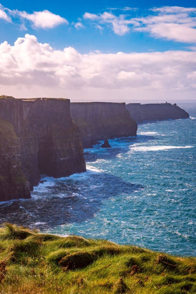 A portrait picture of the Cliffs of Moher on a sunny day with a small section of the foreground showing green grass and the blue sea in the middle and blue skies overhead