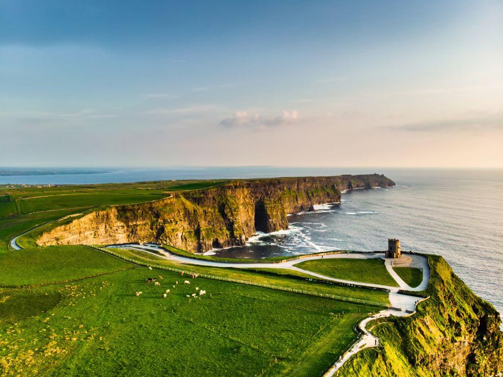 An aerial picture above O'Brien's Tower on the Cliffs of Moher with lush green grass on the left, the sea on the right and blue skies overhead