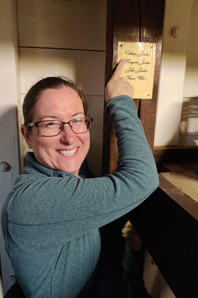 A lady smiling to the camera and pointing to a name on a label at a set of bunks on the Dunbrody Famine Ship