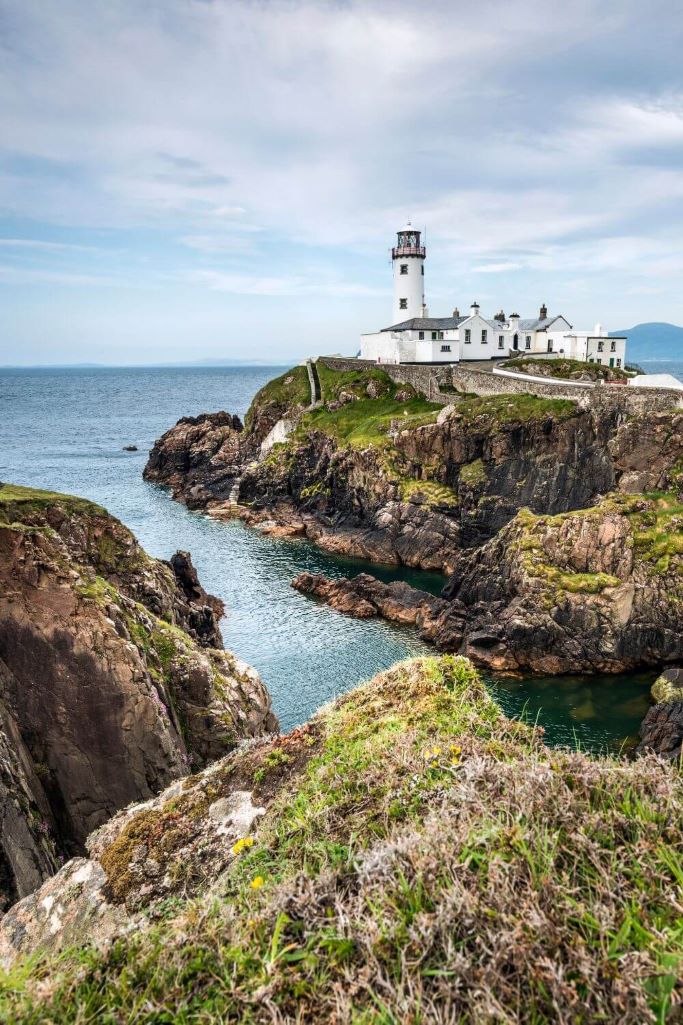 A portrait picture of the Fanad Lighthouse, Donegal with rocky outcrops in the foreground and a cloudy sky above.