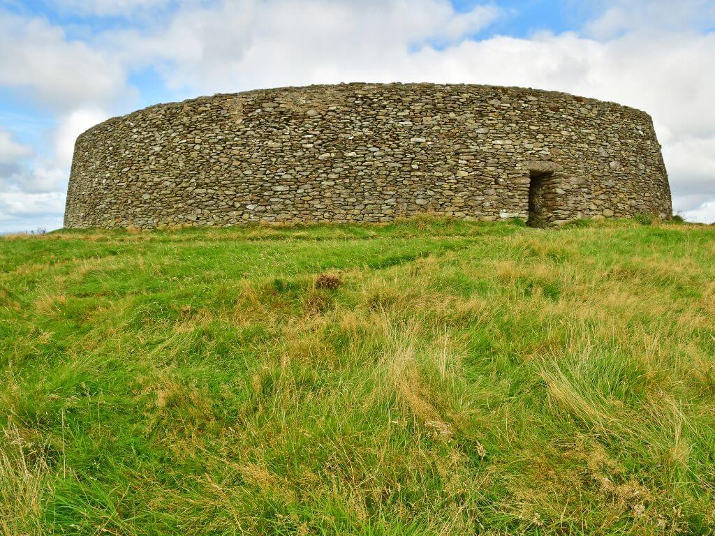 A picture of the entrance doorway and circular stone fort of Grainan Aileach, Donegal with green grass in the foreground and cloudy blue skies overhead