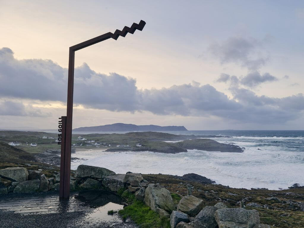 The Wild Atlantic Way sign on the Rosguill Peninsula in County Donegal with waves crashing along the shore and mountains in the background.