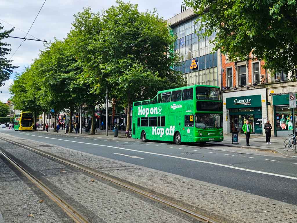 A picture of a green Do Dublin sightseeing hop on hop off bus stopped at a bus stop outside Carroll's gift shop on O'Connell Street in Dublin.