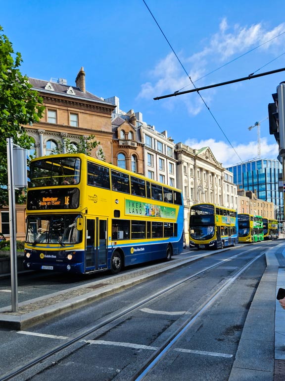 A picture of a row of Dublin buses stopped at traffic lights on College Green with an Luas track beside them.