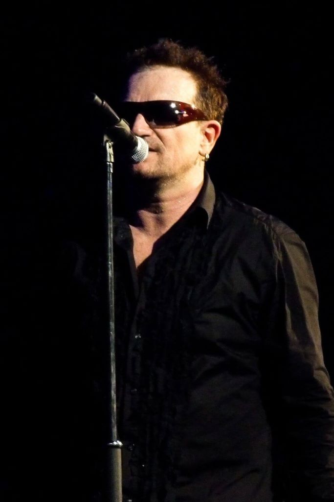 A picture of U2's frontman, Bono standing in front of a microphone with a black background.