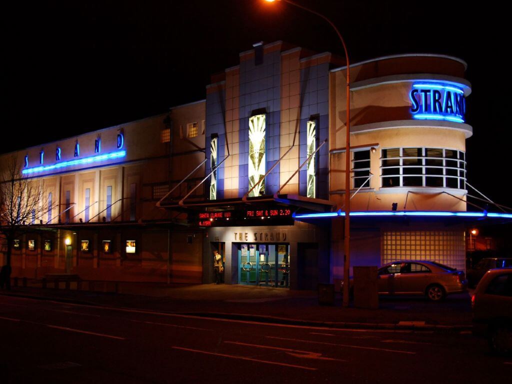 A picture of the exterior of the Strand Cinema in Belfast at night with the name of the cinema illuminated in blue lighting.