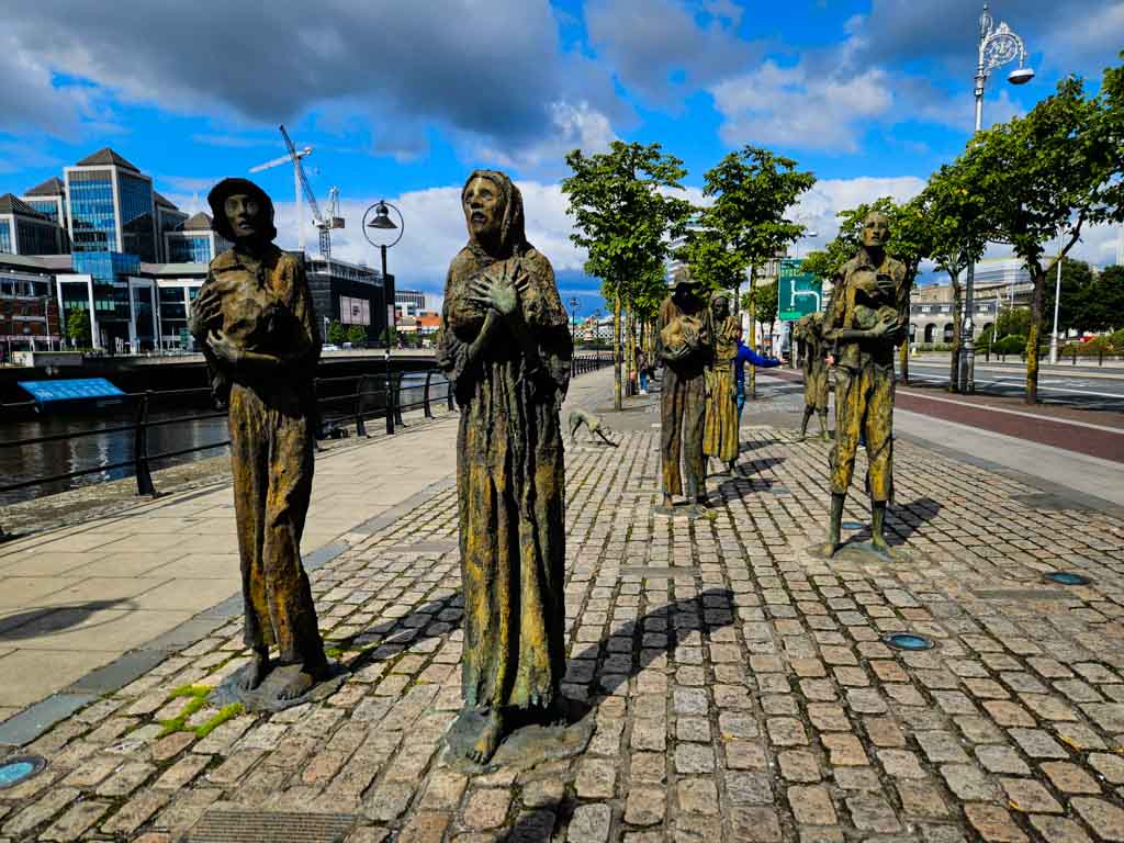 A picture of the Great Famine Memorial which is located on Custom house Quay in Dublin, depicting starving and thin Irish emigrants walking towards a boat to leave Ireland during the Great Famine. There are blue skies overhead and the river Liffey is to the left of the life-sized statues on the quayside.