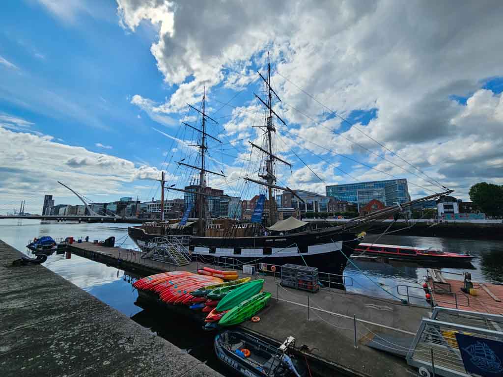A picture of the Jeanie Johnston Famine Ship on the river Liffey from Custom House Quay and blue, cloudy skies overhead with the sun trying to peek around them.