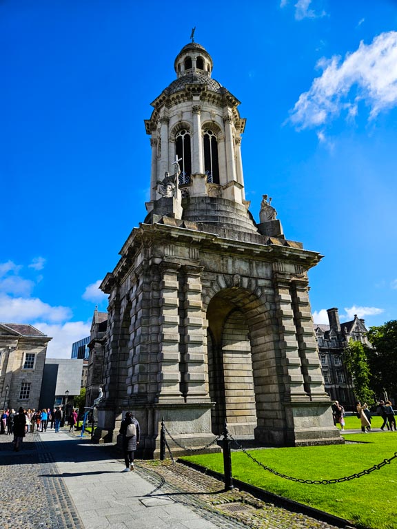 A picture of the Trinity College Campanile in Dublin with blue skies overhead and green grass in the right hand corner of the picture.