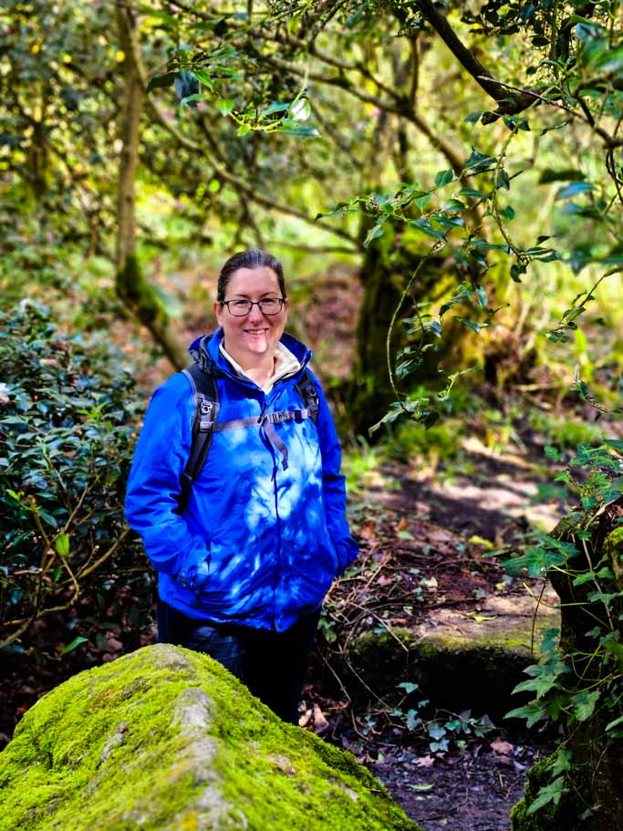 A picture of Cath Jordan from Travel Around Ireland standing in a wooded area with a blue rain jacket on her and smiling to the camera.
