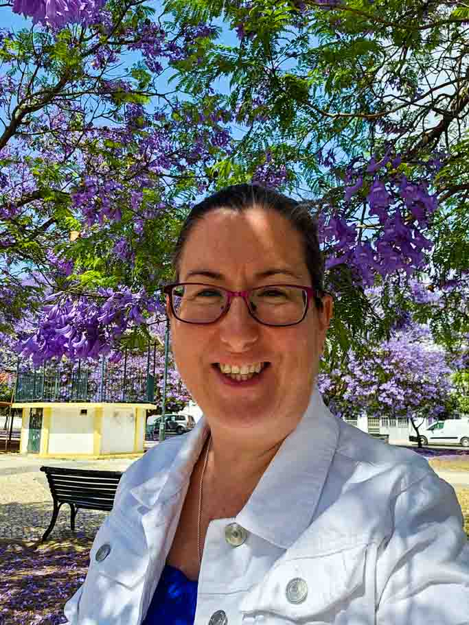 Cath Jordan, founder of Travel Around Ireland, sitting under the purple flowers of a jacaranda tree with a white denim jacket on her and smiling at the camera.