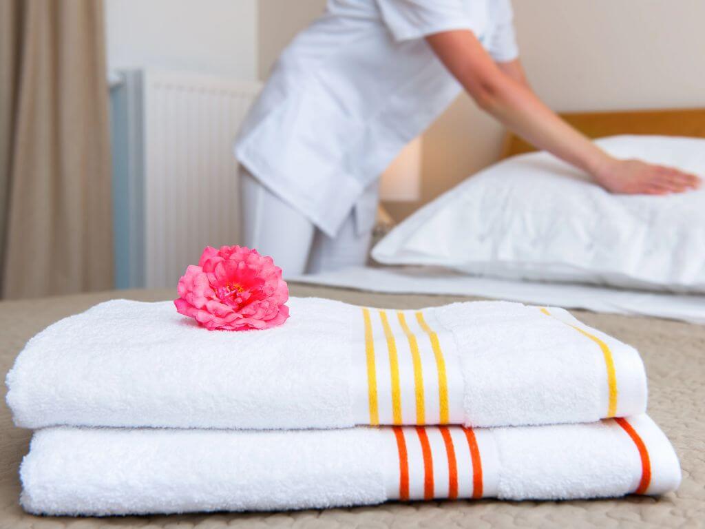 A picture of a hotel room with a housekeeper in the background smoothing out pillows and in the foreground are two folded white towels with a pink flower on top. One towel has yellow stripes and the other has orange stripes.