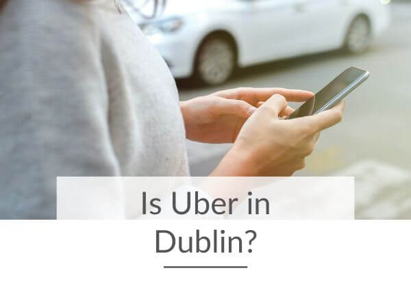 A picture of a person in a grey top using a cell phone and in the background a white car is visible. There is text overlay across the lower half of the picture saying is Uber in Dublin?