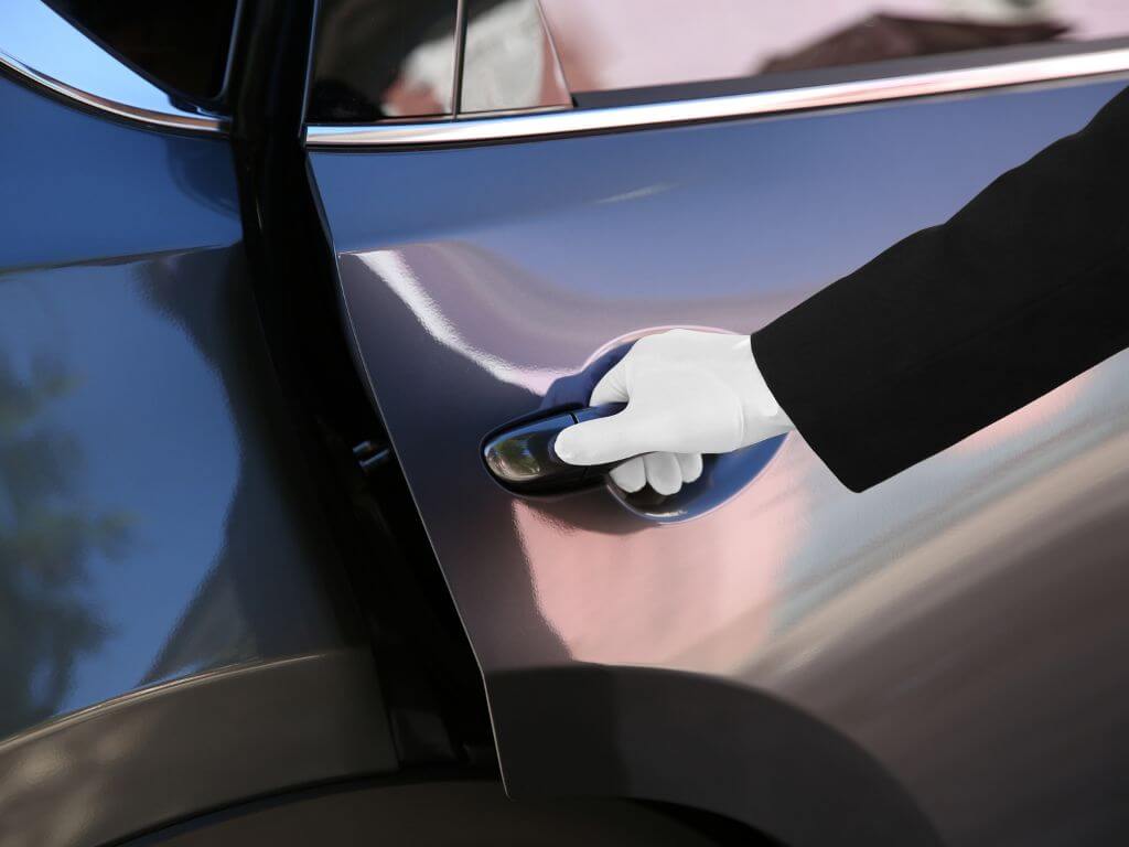 A picture of a white gloved hand opening a car door. The car is black and the person with the gloved hand is wearing a black suit jacket.
